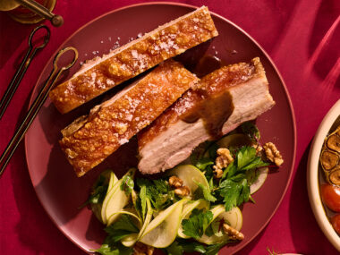 Twice-cooked pork belly with pickled apple salad and maple-glazed cabbage on red background.