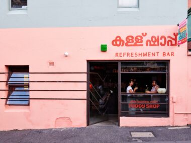 The exterior of a restaurant with a pink wall. Two people are sitting in the window.