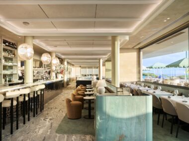 A photo of a large dining room. On the left-hand side there is a bar, and on the right there is a window with a view of the ocean.